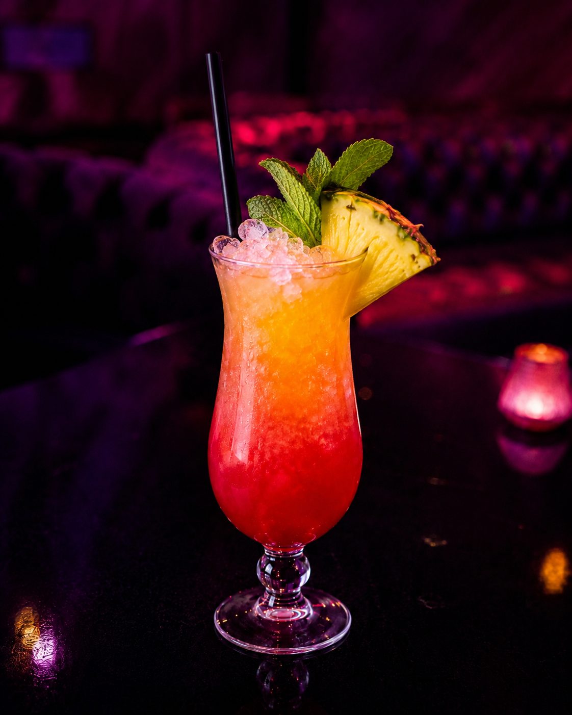 Orangeroter Cocktail im Hurricane-Glas mit Ananasscheibe und frischer Minze.